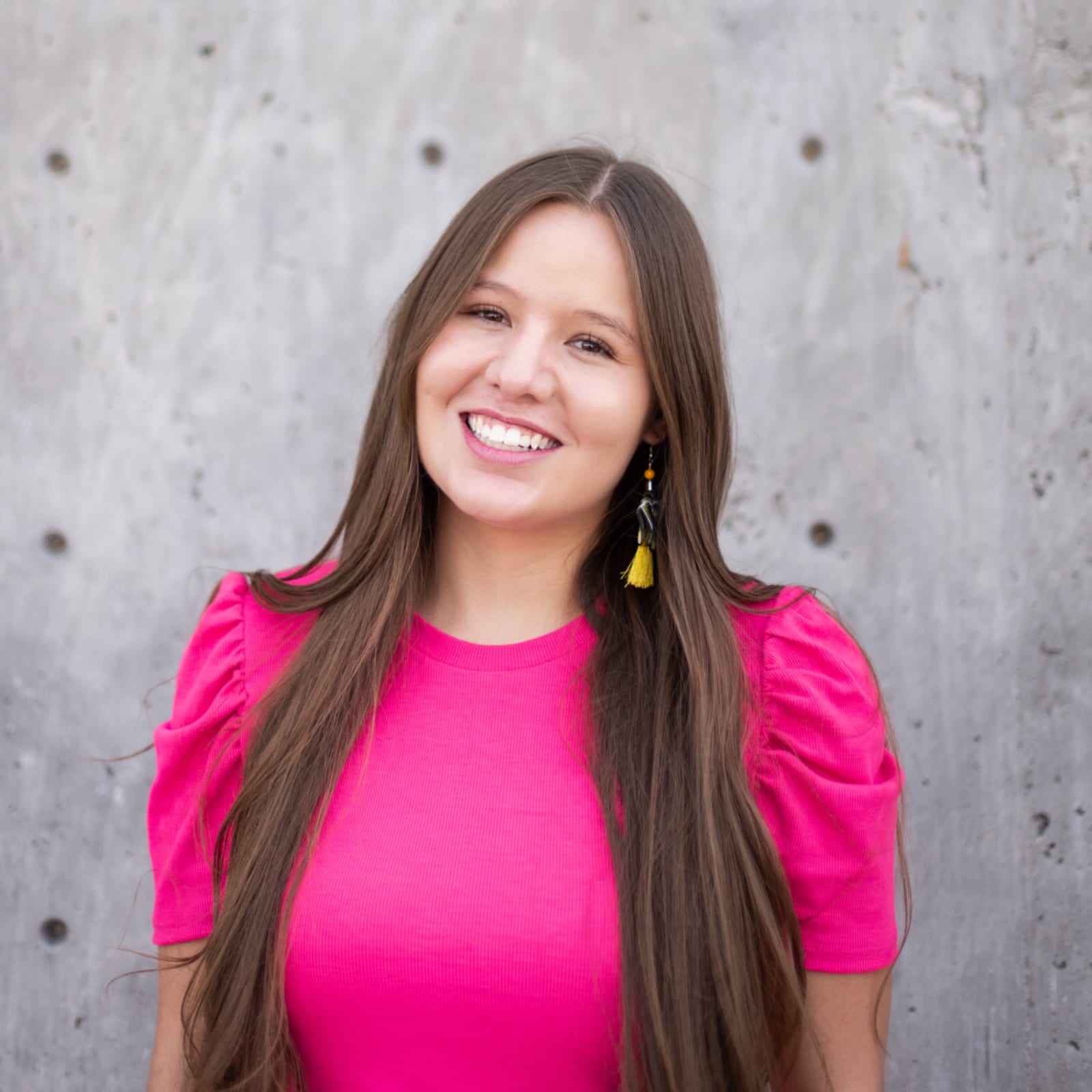 Alissia smiling in a pink top and a yellow tassel earring, against a concrete wall